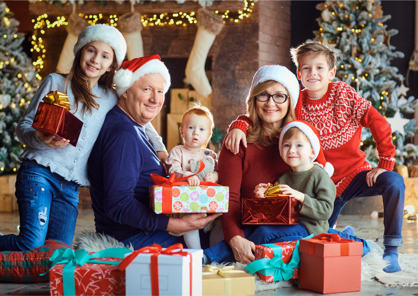 Multigenerational family in Santa hats exchanging wrapped presents in front of a glowing Christmas tree and fireplace, celebrating together in a cozy holiday living room.