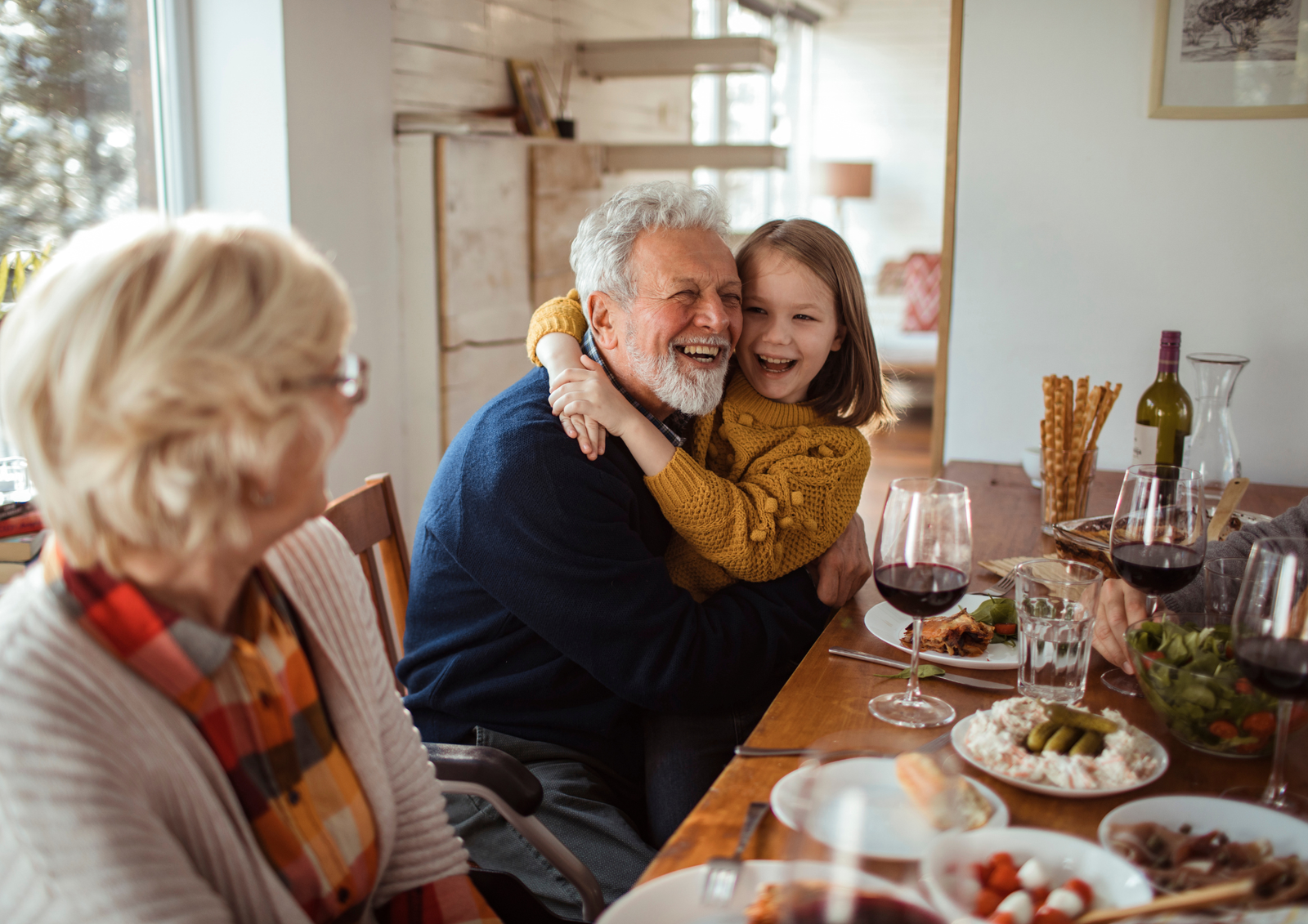 A young girl smiling and hugging her grandfather around the neck at a festive holiday dinner table with family.
