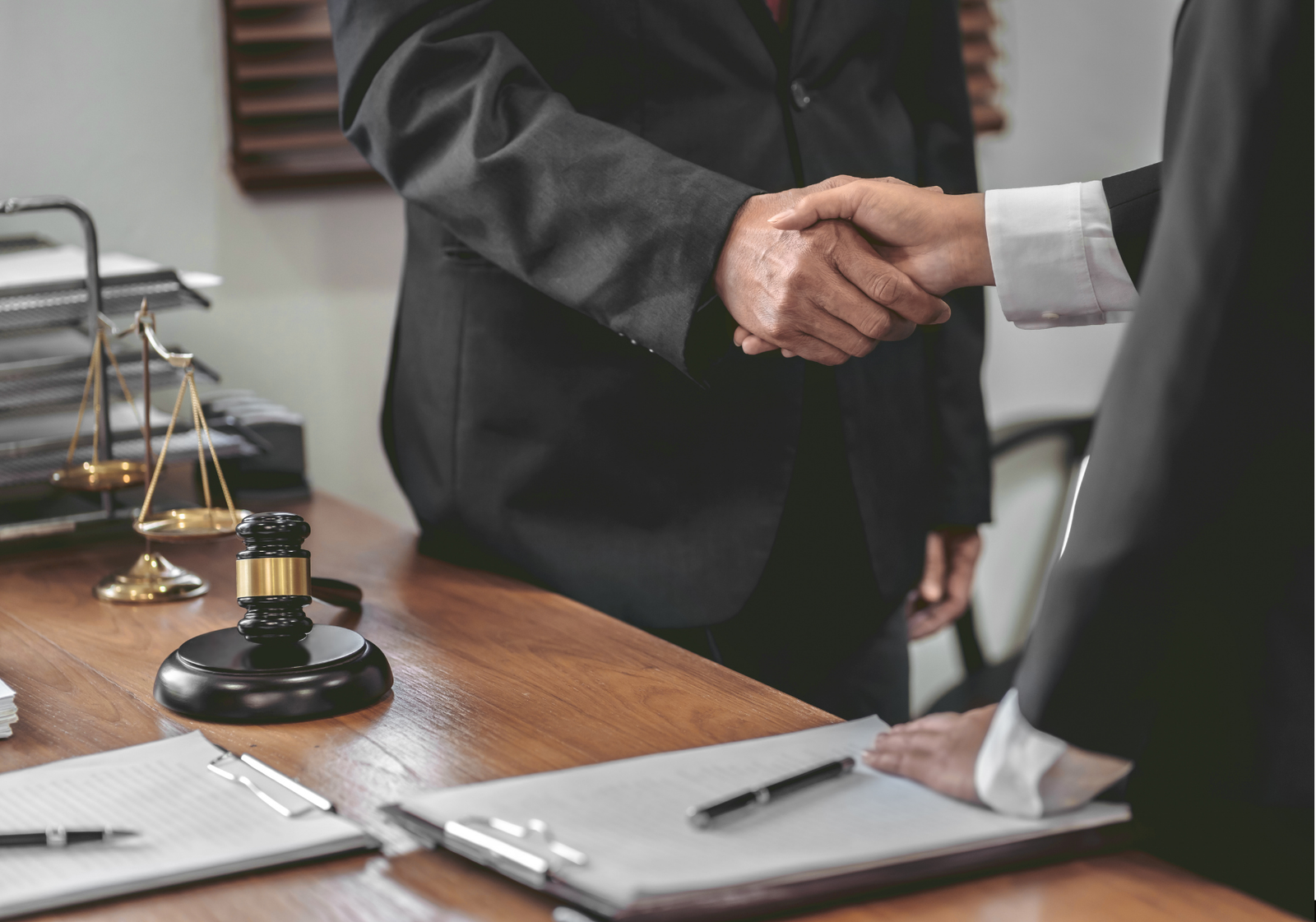 Two attorneys shaking hands across a desk with a gavel, legal scales, and documents during a client meeting.