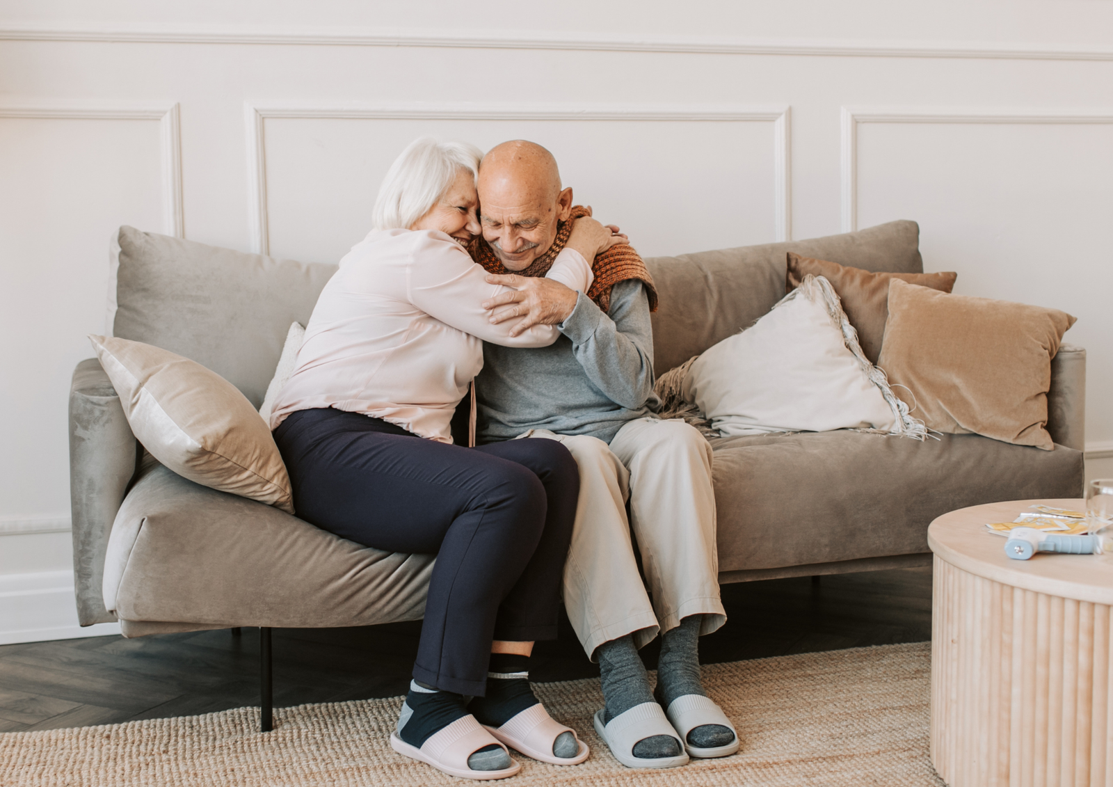 An elderly woman warmly hugging an older man as they sit together on a cozy living room sofa.