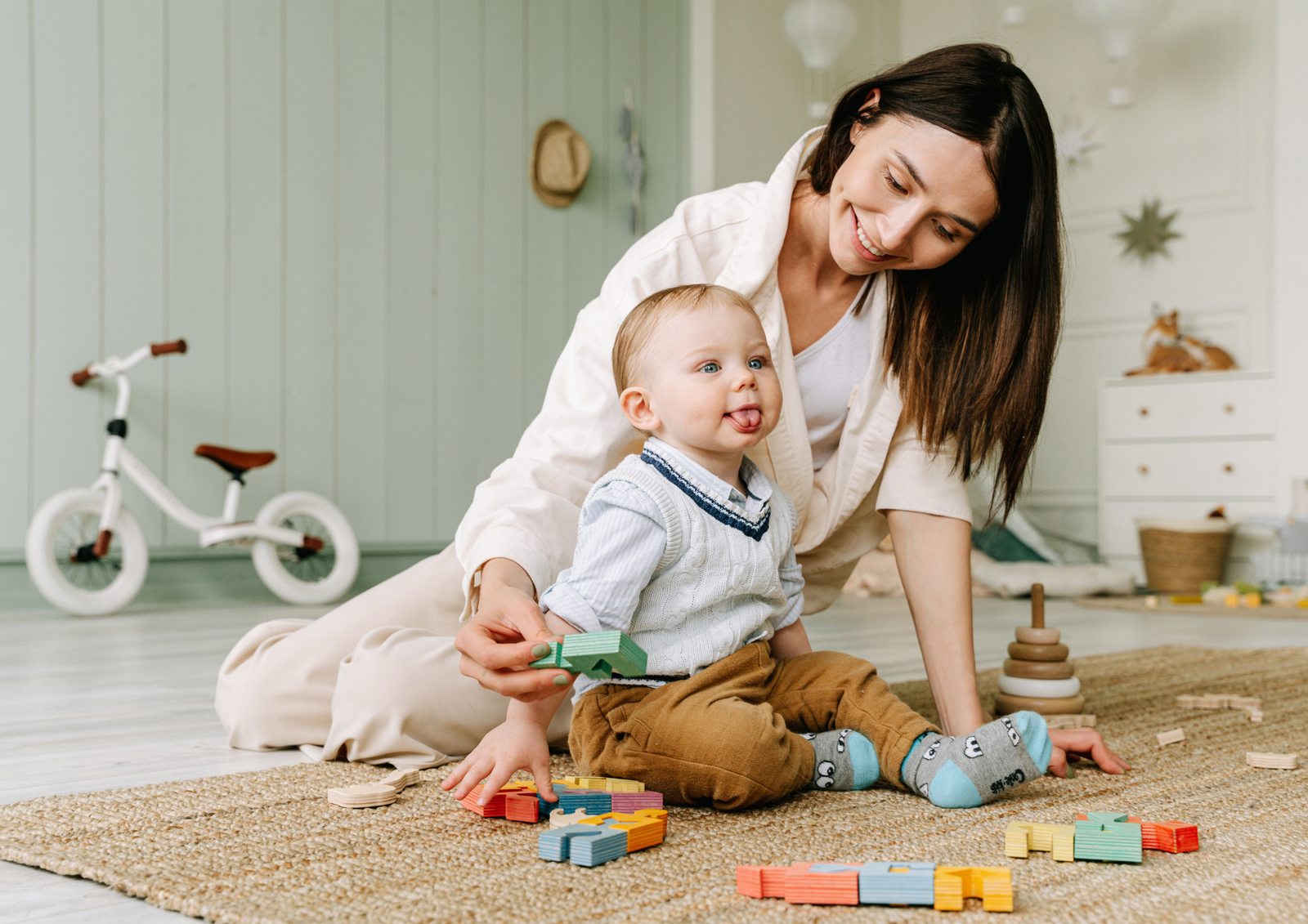 A smiling mother playing with colorful wooden building blocks on a rug alongside her toddler in a playroom.