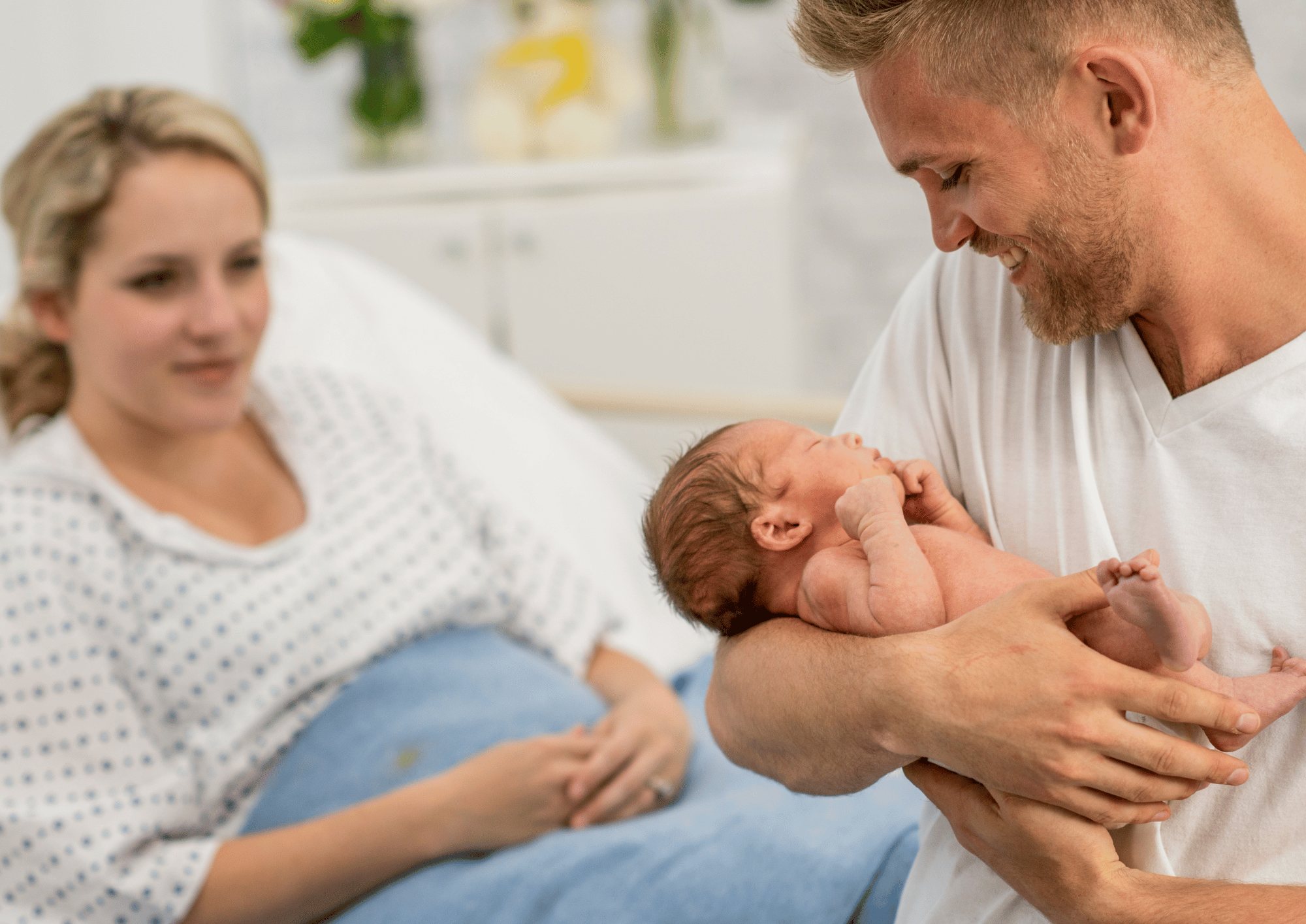 Best Gift Baskets for New Dads: A smiling father cradling his newborn baby in a hospital room beside his partner.