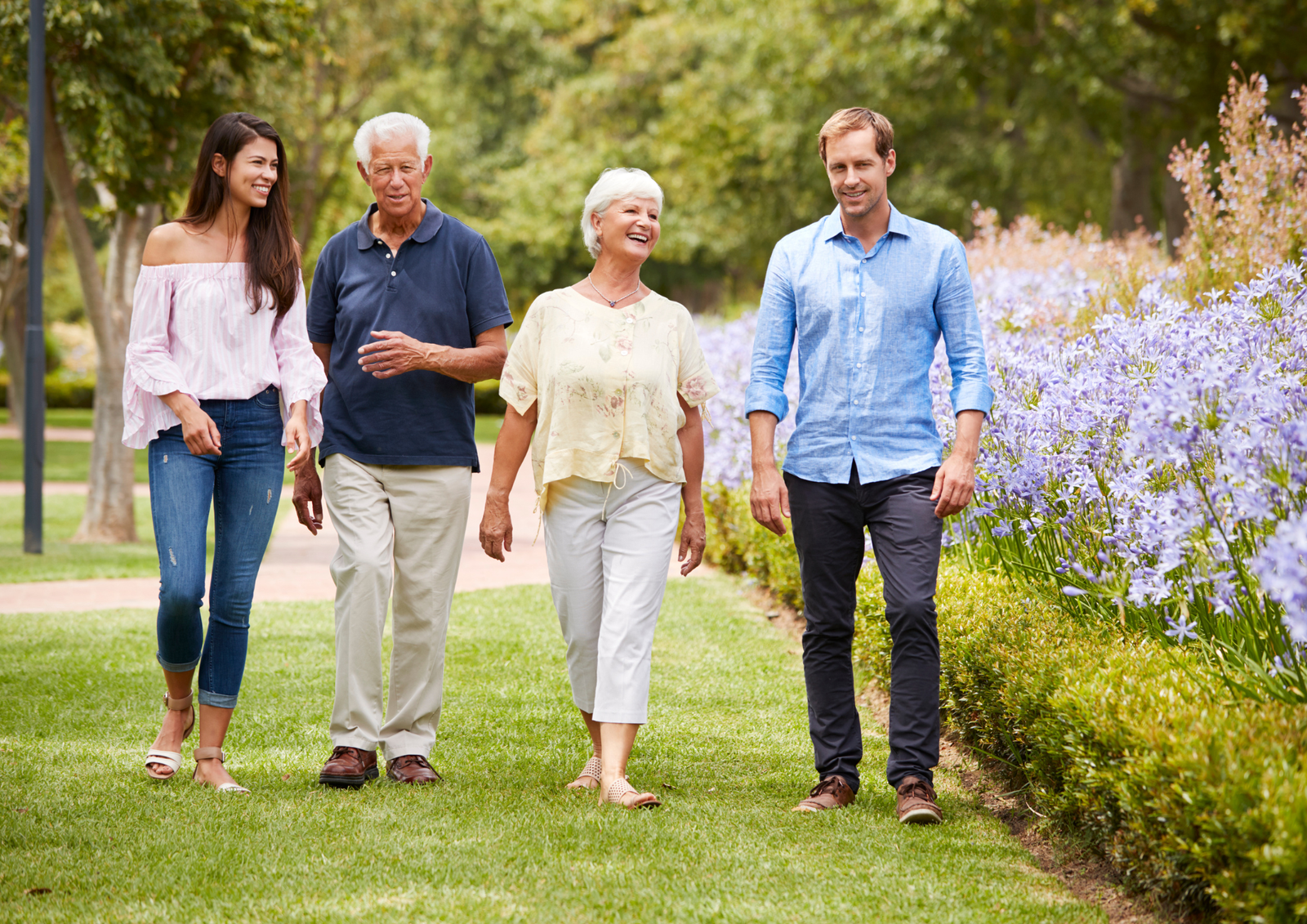 Two couples smiling and walking together along a garden path with purple flowers.
