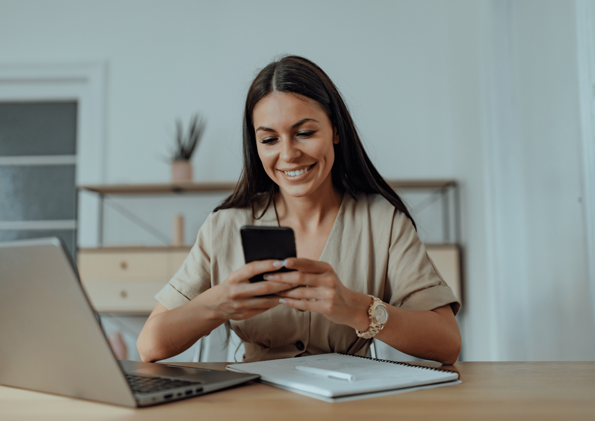 A smiling woman sitting at a desk with a laptop and notebook while checking her smartphone to send a text message.