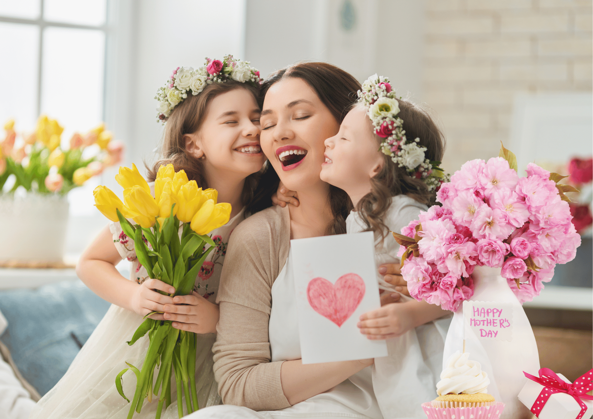 A mother laughing while receiving Mother’s Day gifts including flowers, and a greeting card from her daughters.