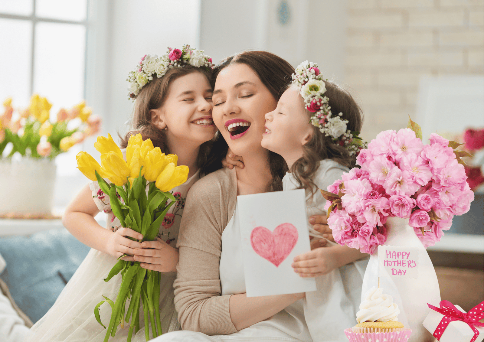 A mother laughing while receiving Mother’s Day gifts including flowers, and a greeting card from her daughters.