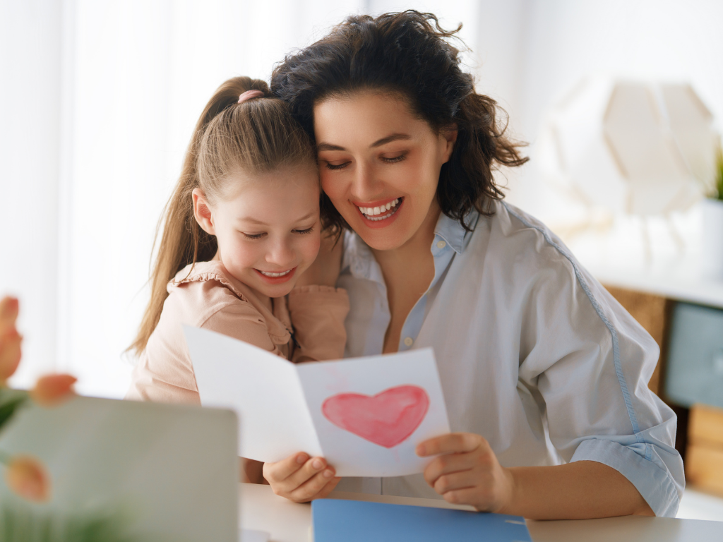 A smiling mother and daughter look at a handmade Mother's Day greeting card featuring a large pink heart.