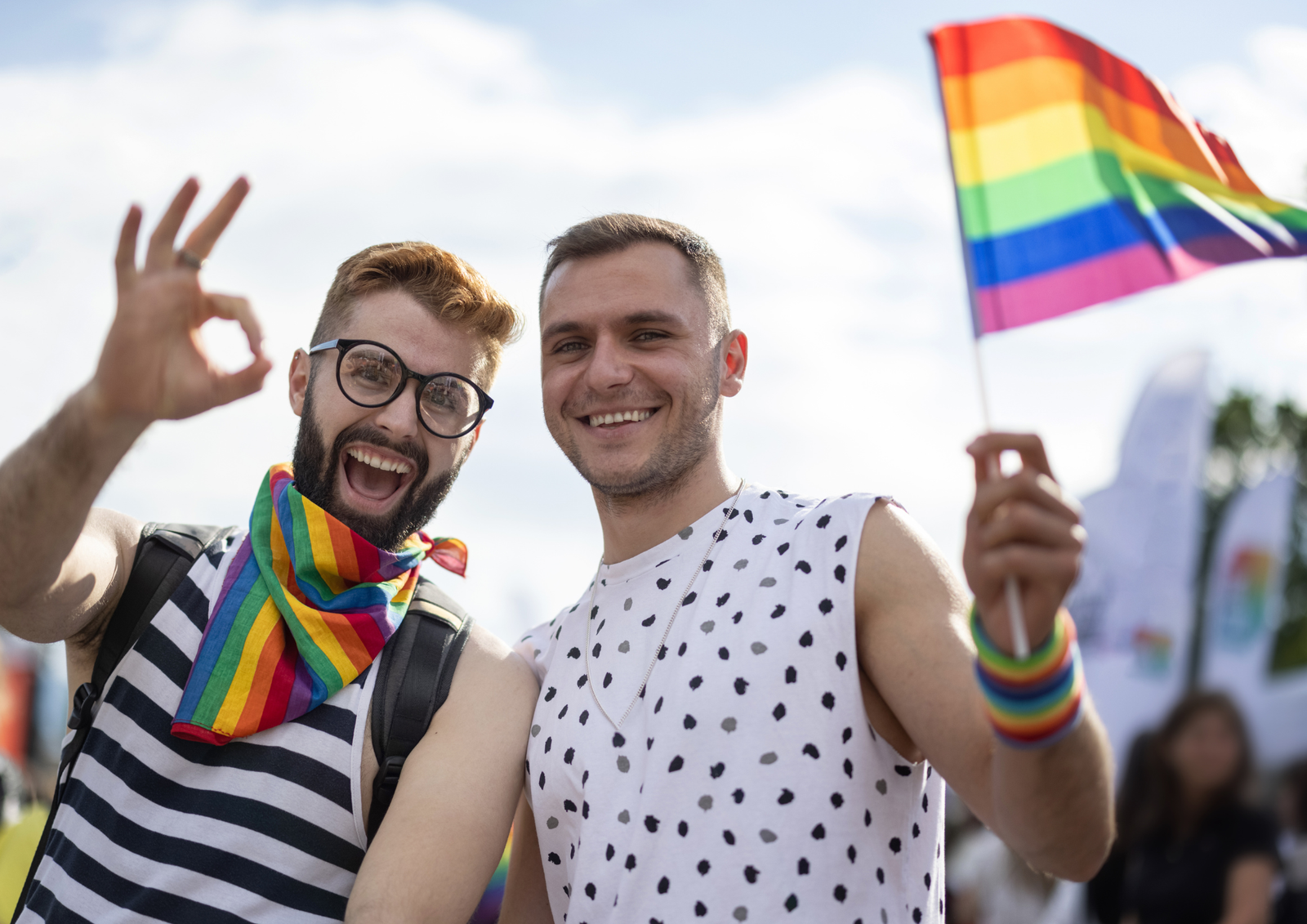 Two smiling men celebrating at an outdoor Pride festival, one holding a rainbow flag and the other making an OK hand sign.