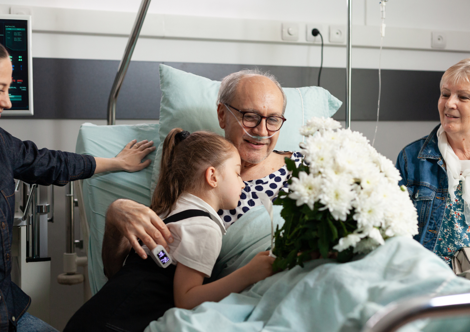 A young girl hugging her grandfather as she presents him with a bouquet of white flowers in his hospital bed after surgery.