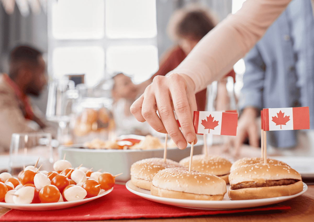 Canada Day Gift Baskets: A hand placing a tiny Canadian flag toothpick into sliders on a party table with appetizers.