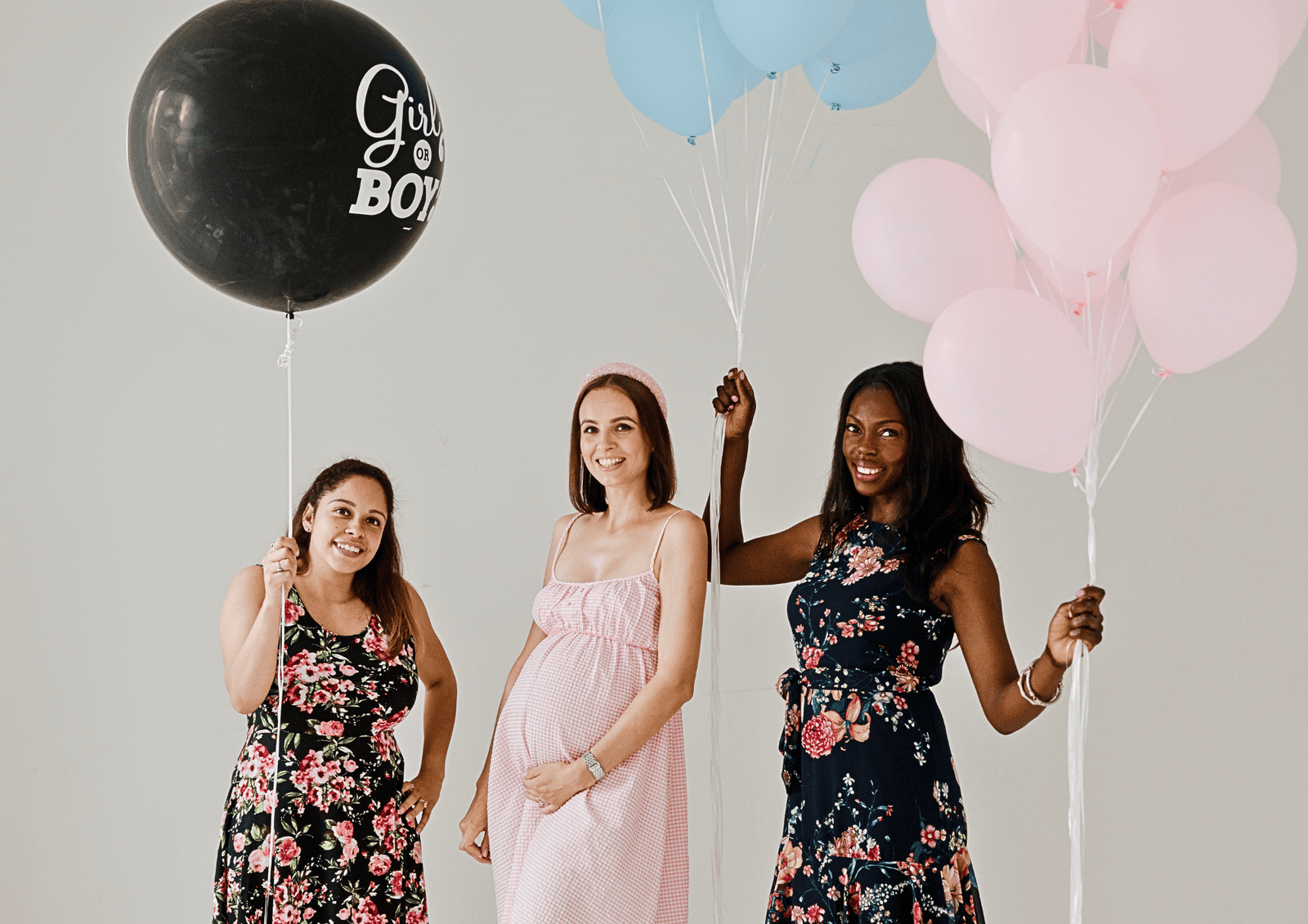 Three smiling women holding pink, blue, and a large black "Girl or Boy" reveal balloon at a baby shower party.