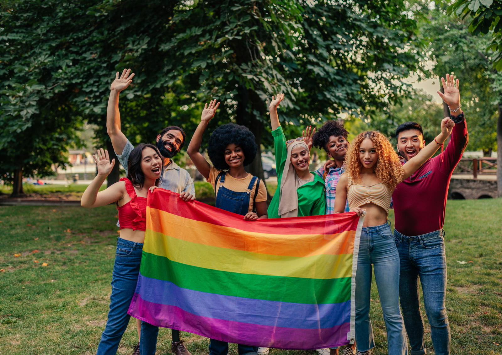 A diverse group of friends smiling and waving while holding a rainbow Pride flag together at an outdoor park party.