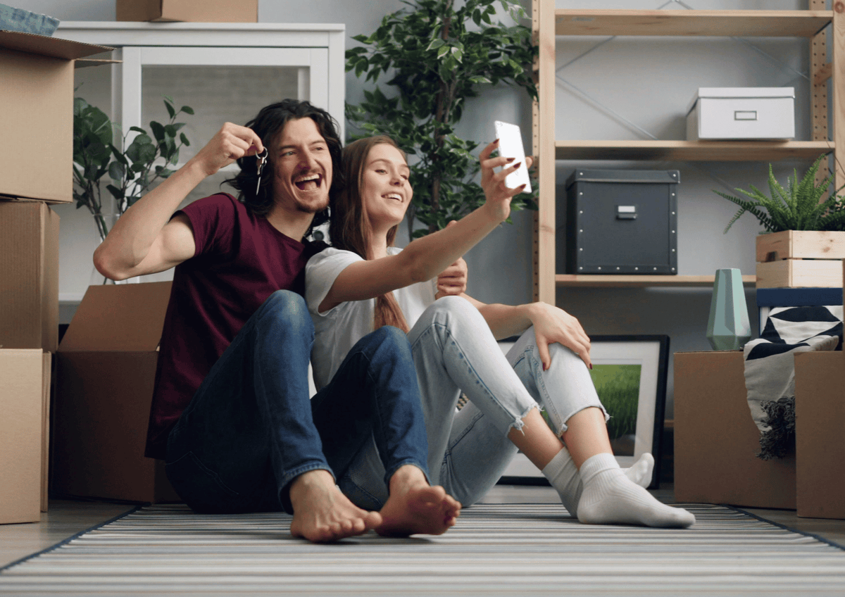 A happy couple sitting on the floor of their new home taking a selfie while holding up a pair of house keys.