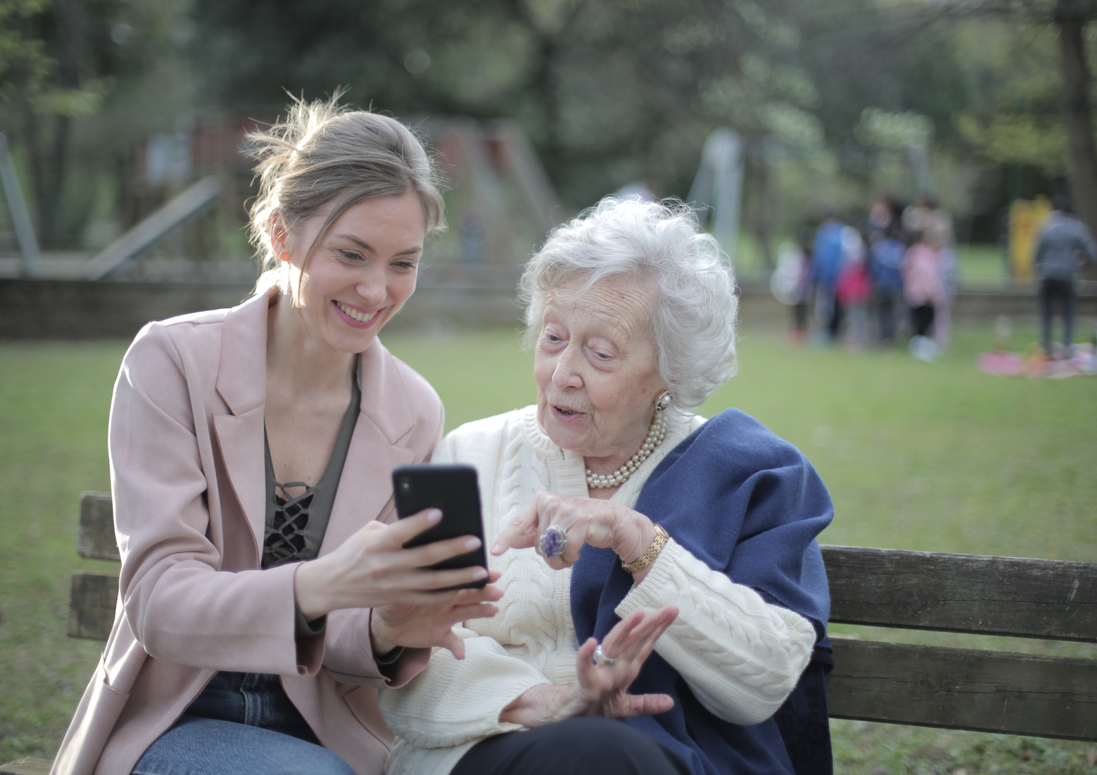 Young woman and elderly woman sitting on a park bench looking at a phone together.