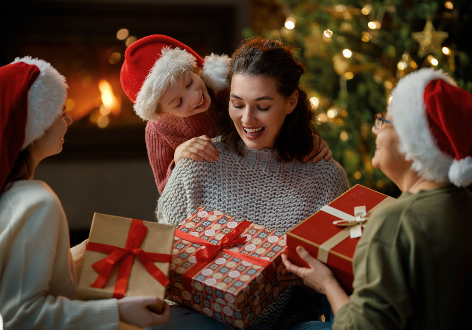 Smiling woman receiving Christmas gift boxes from family members wearing Santa hats by the fireplace.