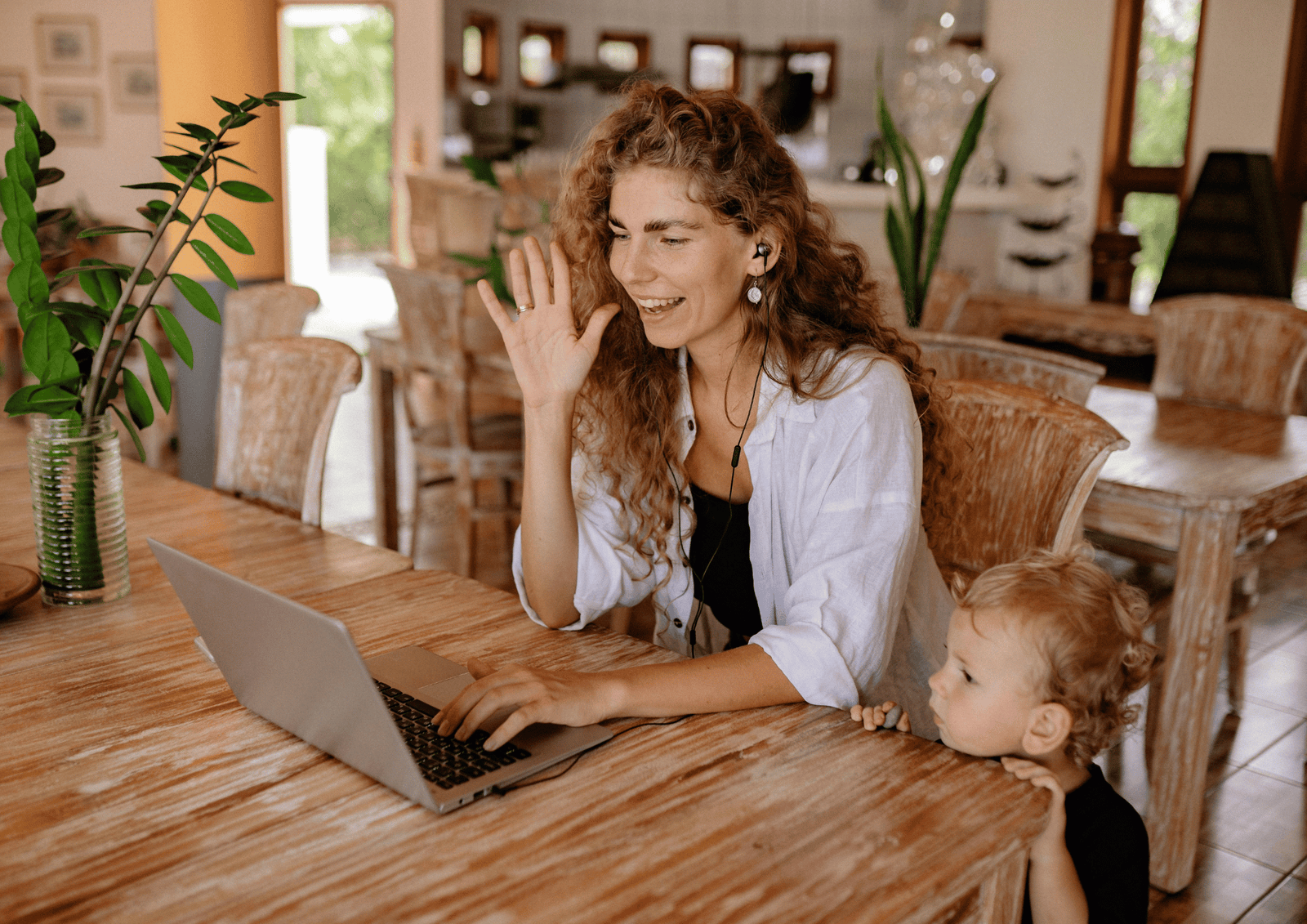A smiling woman with a toddler waving during a video call on her laptop at a wooden dining table.