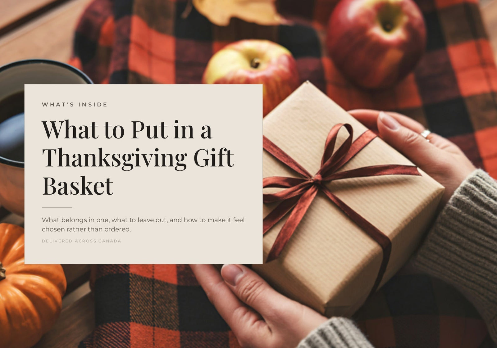 Hands holding a wrapped gift box tied with a red ribbon on a plaid blanket with apples and a pumpkin.